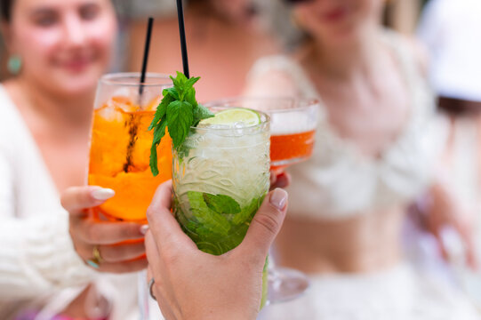 Friends toast with cocktails including a mojito, spritz, and coupe drink in Corfu, Greece. A close-up shot highlights hands holding iced glasses at an outdoor table.