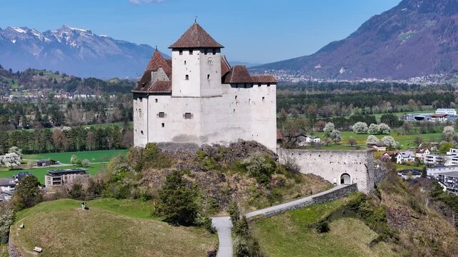 town rooftops solar panels blossom trees gutenberg castle hilltop snowcapped mountain ridge backdrop path uphill balzers liechtenstein rhine valley farmland drone footage aerial orbit heritage site