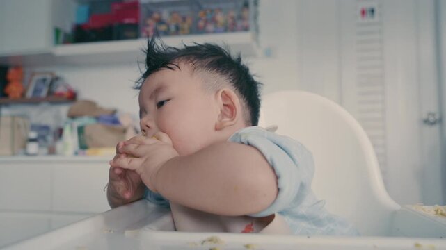 Asian baby boy eating boiled corn on high chair, BLW (baby-led weaning) concept, healthy solid food introducing to infant at home