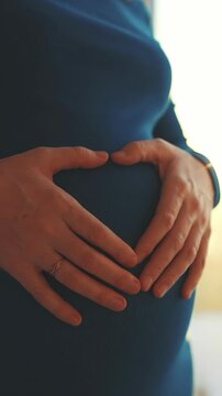 A woman gently holds her pregnant belly, forming a heart shape with her hands. The soft light creates a warm atmosphere around her and her belly.