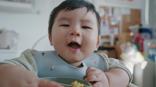 Asian baby boy eating boiled corn on high chair, BLW (baby-led weaning) concept, healthy solid food introducing to infant at home