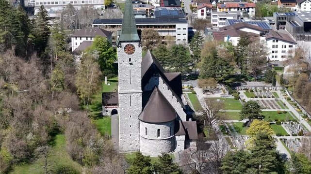 bell tower clock face pyramidal spire parish church close aerial orbit cemetery graveyard dense town rooftops solar panels forested hillside balzers liechtenstein heritage site drone footage