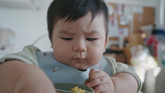 Asian baby boy eating boiled corn on high chair, BLW (baby-led weaning) concept, healthy solid food introducing to infant at home