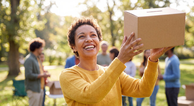 Smiling woman holding donation box photo at outdoor charity event with volunteers