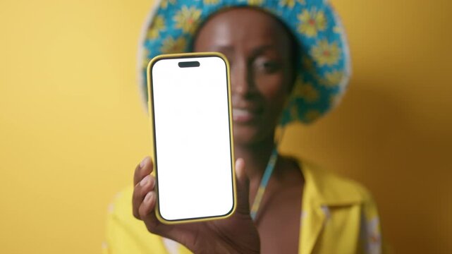 Stylish young black woman is posing with her smartphone with blank white screen against yellow background. She is wearing bob hat and blouse. 