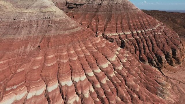 Aerial view sweeping over dramatic red and white striped sandstone formations with deep erosion patterns under blue sky in Aktau Mountains, Altyn Emel National Park, Kazakhstan