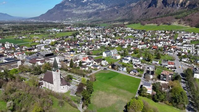 steep rock face mountain flank towers balzers valley town castle hilltop parish church spire green farmland fields snowcapped alpine peaks aerial panorama liechtenstein drone footage heritage site