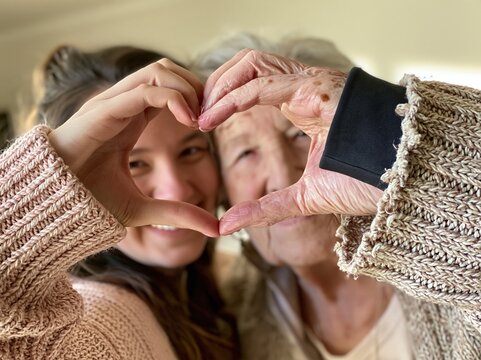 Hands forming a heart shape by a grandmother and granddaughter, smiling in a soft light interior for family care and love