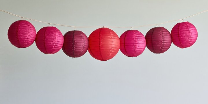 Seven round paper lanterns hanging on a curved cord with vivid fuchsia and red tones against a plain white background for party decorations