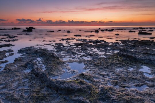 Tranquil rocky shoreline with shallow tide pools reflecting pastel dawn colors over a calm ocean. Ideal background for wellness, travel, and nature conservation themes.