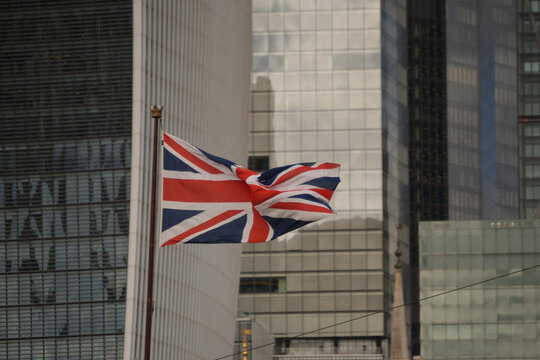 British flag waving Union Jack symbol, fluttering in wind, flagpole, urban setting, modern skyscrapers background, corporate glass buildings, London financial district, daylight