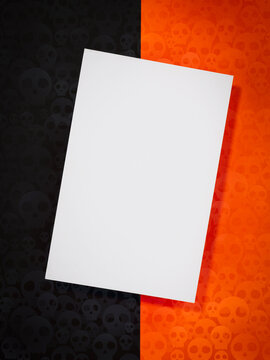 Blank white greeting card rests on an orange and black skull-pattern background, creating a Halloween invitation mockup for event announcements, seasonal promotions, party messages and custom designs.