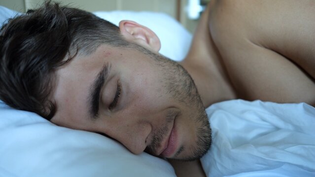 Portrait of handsome man sleeping peacefully in bed in modern hotel room or home interior. Young guy resting under soft linen in cozy indoor atmosphere with natural comfort and relaxation