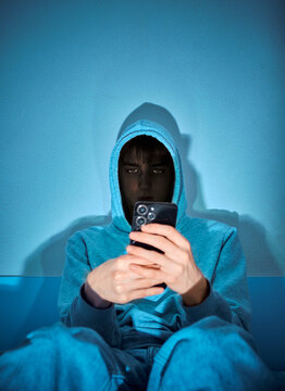 Hooded person using a smartphone in dramatic blue lighting, creating a mysterious cybercrime and online privacy concept with strong shadows and a dark, technological atmosphere