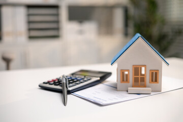 A miniature house stands on paperwork beside a calculator and pen. This still life illustrates mortgage calculations, home buying budgets and property insurance, with copy space on the left. © DK_STUDIO
