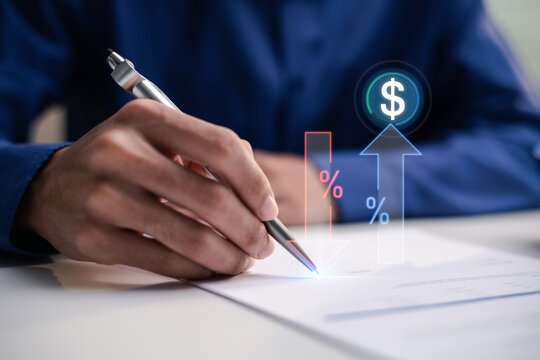 A person writes on a document beneath financial graphics at a desk. Up and down arrows, percentage signs and a dollar symbol illustrate interest rate changes, borrowing costs and financial planning.