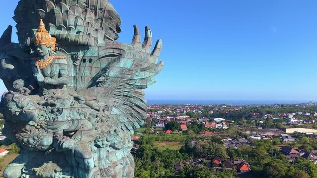 Filmed from above and behind, the broad bronze wings of Garuda spread wide beneath the figure of Vishnu, with the residential sprawl and coastline of southern Bali stretching to every horizon below