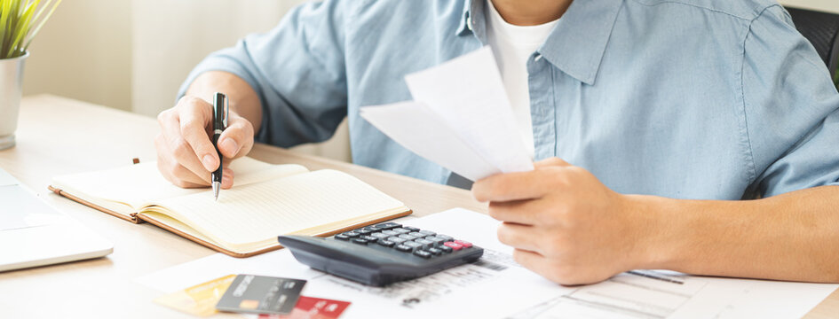 Stressed asian young businessman hand write management budget for saving cost expenses of monthly, male holding bills and receipt for to payment on table at home. Financial, finance of banking concept