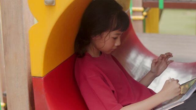 Asian kid drawing digital art on tablet inside a playground tunnel. Selective focus highlights the creative elementary student enjoying casual hobby and relaxing time outdoors with technology.
