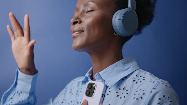Young Black American woman dancing agains blue studio background in headphones and with smartphone and smiling