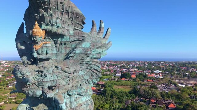 Flying alongside the GWK monument, the camera reveals the massive scale of Garuda's bronze wing against the white stone podium building, with the vast urban and coastal panorama of southern Bali beyon