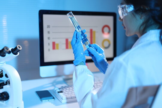 Scientist analyzing chemical liquid in laboratory with microscope. Scientist wearing protective glasses and gloves, holding a test tube with liquid, looking at a computer monitor with data charts.
