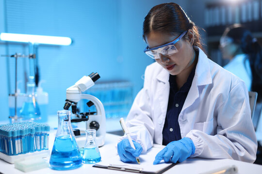 Female scientist conducting research, writing notes in laboratory, working with chemicals and microscope, recording experiment results.