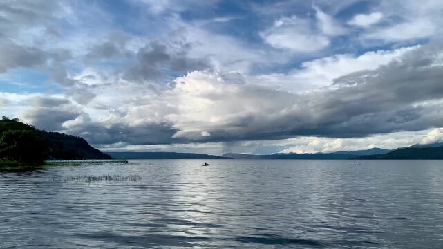 Wide view of a local fisherman paddling a small boat across the quiet waters of Lake Toba in North Sumatera, Indonesia. Ideal commercial background for travel and lifestyle.