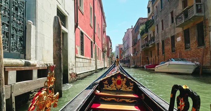 Venice, Italy - August 23, 2026: POV shot from a traditional carved gondola moving through a calm narrow Venetian canal lined with weathered historic buildings and moored motorboats on a sunny summer 