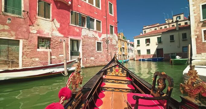POV shot from a luxury carved gondola floating along a sunlit Venetian canal junction flanked by weathered rustic red brick houses and moored boats.