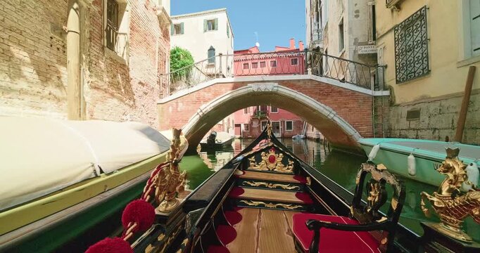 POV shot from a traditional carved gondola floating along a narrow Venetian canal directly towards the picturesque brick Ponte Colombo bridge on a bright sunny summer 