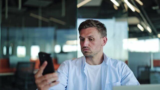 Frustrated bearded businessman in blue shirt waving smartphone in air while experiencing poor mobile connection, searching for cellular network signal or Wi-Fi in modern office.