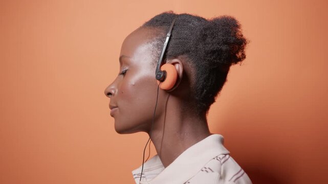 Young beautiful Black American woman is dancing against orange studio background with copyspace as she is enjoying music in vintage headphones with eyes closed. 