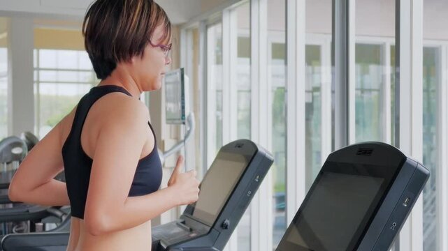 Active Asian woman in her forties wearing glasses and a black sports bra runs on a treadmill inside a modern brightly lit gym. She is focused on her cardio workout to maintain a healthy lifestyle.