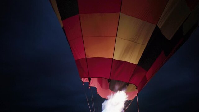 Propane burner flame firing into mouth of checkered hot air balloon envelope during night inflation process, bright fire nozzle illuminating colorful fabric against dark sky, close up low angle shot
