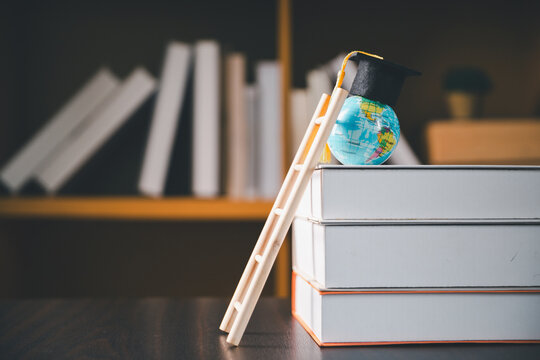 Mini world globe wearing a graduation cap sits atop stacked books beside a wooden ladder in a library, symbolizing education, academic progress, global learning, achievement, ambition, and success.