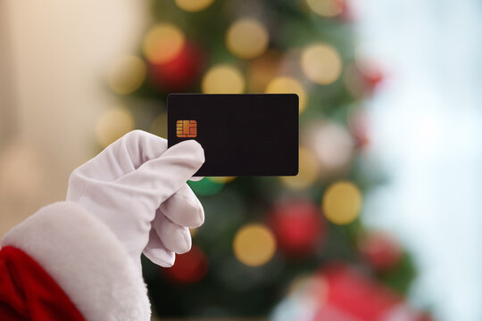 Santa Claus holding credit card in front of  Christmas tree, representing holiday shopping, seasonal spending, cashless payment, discounts and Black Friday sales.
