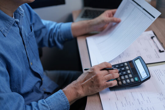 Man using a calculator while reviewing financial documents and bills at home, representing accounting, expense analysis, budgeting, monthly payments, debt and personal finance planning.