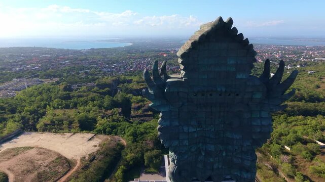 A close lateral drone pass along the upper section of the GWK statue reveals the intricate facial features of Vishnu's crowned head and the feathered detail of Garuda's neck and wings against the open
