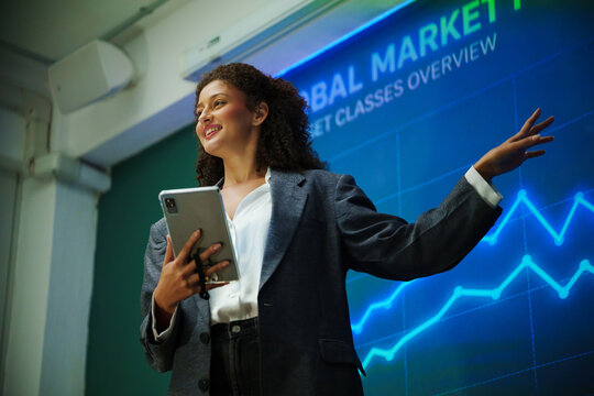 Confident businesswoman giving a financial market presentation with digital graphs on screen, representing leadership, finance analysis, business strategy, performance and professional speaking.
