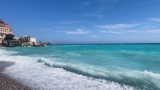 Blue waves crash against the shore, creating a vibrant and dynamic seascape. The bright sky and deep blue sea blend beautifully of the Mediterranean Sea near Nice, Cote d'Azur, southern France.
