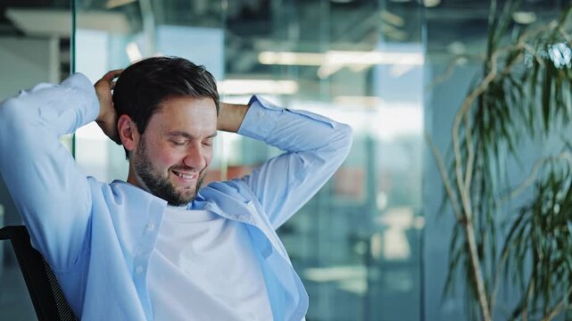 Businessman in a light blue shirt and white t-shirt leans back, stretching his arms behind his head, smiling contentedly in a modern office environment. He looks relaxed and satisfied after work.