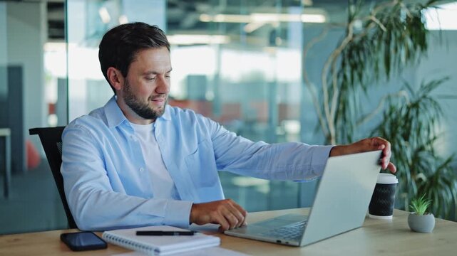 Businessman in a light blue shirt and white t-shirt leans back, stretching his arms behind his head, smiling contentedly in a modern office environment. He looks relaxed and satisfied after work.