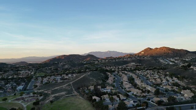 Aerial back from hills and over residential neighborhoods and streets in Corona, California during sunset. Riverside County, in the Inland Empire near Los Angeles. Shot during January, 2024.