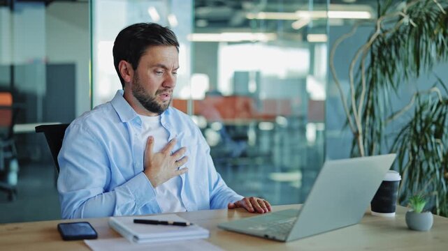 Businessman in a light blue shirt experiences sudden chest pain while typing on his laptop at a desk in a modern office, then recovers. He appears stressed and concerned about his health.