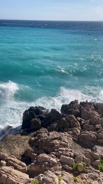 Blues waves crash against the rocks, creating a vivid and energetic coastal scene. The bright sky and deep blue sea blend beautifully of the Mediterranean Sea near Nice, Cote d'Azur, southern France.
