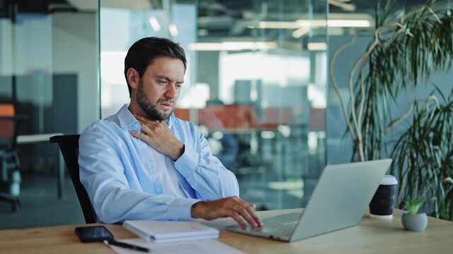 Businessman in a blue shirt experiences throat pain while typing on his laptop at a modern office desk, indicating illness or discomfort.