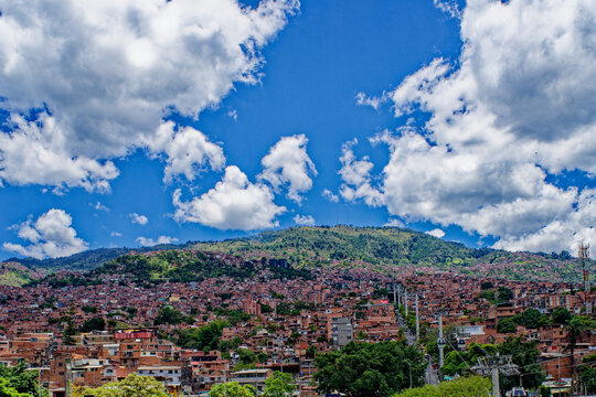 Low-angle view of Medellin, Colombia, showing dense brick houses on the hillside and Metrocable transit lines scaling the mountain under a bright, cloud-filled blue sky.