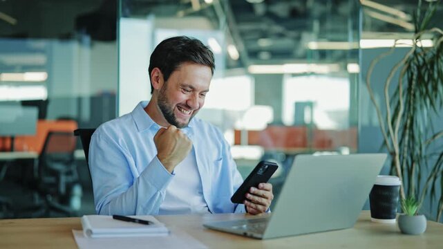 Corporate analyst celebrates an online achievement, fist-pumping with joy and a wide smile while using a smartphone at his modern office desk.