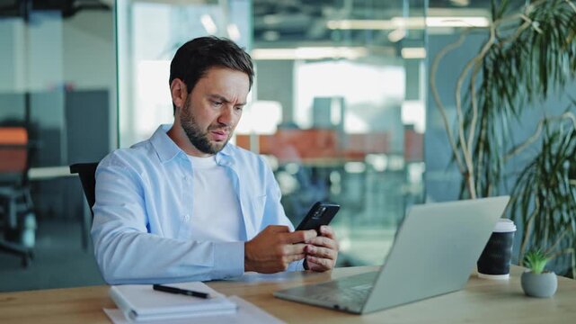 Corporate analyst, dressed in light blue shirt, shows a look of concern while operating his smartphone at an office desk. He appears to be reacting to bad news or a stressful message on mobile device.